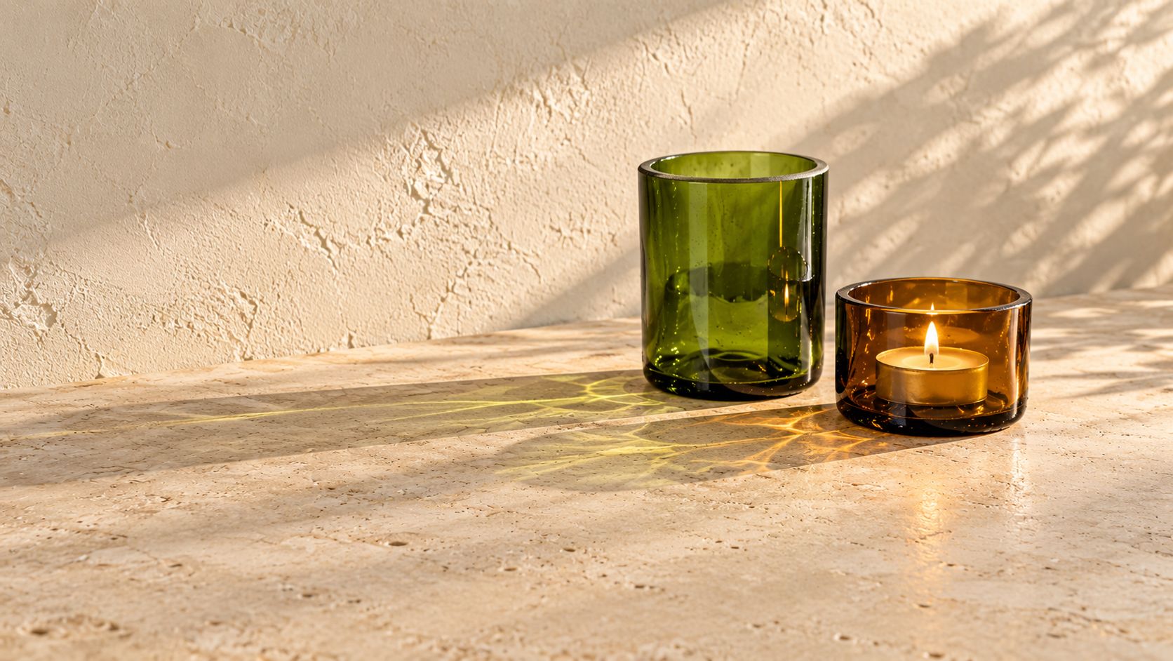 Två föremål gjorda av avkapade flaskbottnar, på en stenskiva i solljus. Det högre är i grönt glas och tomt, det lägre i brunt glas med ett tänt värmeljus. Glasen kastar grönt och gyllene ljus på stenen.